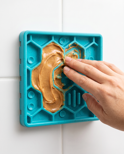 Hand spreading peanut butter into the Joobello silicone lick mat maze grooves suctioned to a tile wall